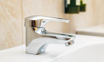 A modern stainless steel faucet is located on a sink. In the background, bottle-shaped care products are visible.