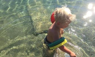 A little boy with a swimming ring is playing in the clear water. The sun reflects beautifully on the surface of the water.