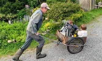 A man is pulling a cart on a gravel path, in which two small children are sitting. The surroundings consist of green plants and bushes.