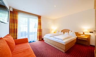 A bright hotel room with a large bed and a cozy sofa. The room has modern amenities and a balcony with an outdoor view.