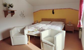 A cozy room with a double bed and two white armchairs. The walls are painted yellow, and there is a small table between the armchairs.