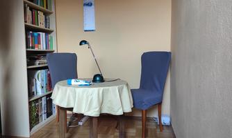 A small room with a round table and two chairs. There is a bookshelf on the wall and a lamp is on the table.