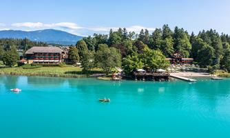 A picturesque landscape by the water with a green shore and a blue, clear lake surface. In the background, trees and a beautiful building are visible.