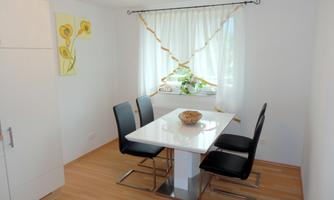 A bright dining area with a white table and black chairs. A window with curtains and a plant illuminates the room.