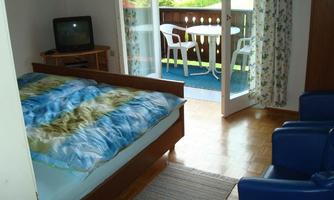 A cozy bedroom with a double bed and a television. Through the door, a balcony with chairs and a table is visible.