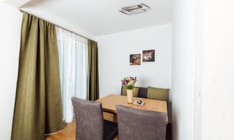 A cozy dining area with a wooden table and four chairs. The windows are decorated with green curtains and a bouquet of flowers stands in the center.