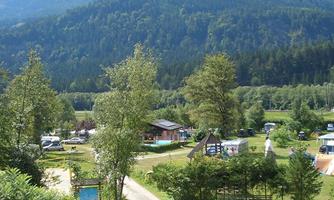 A picturesque landscape with green trees and mountains in the background. In the foreground, there is a campsite with caravans and a playground.