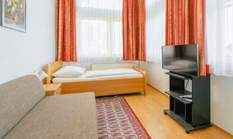 A cozy room with a bed, a couch, and a television. The windows are adorned with light curtains and the floor is covered with a carpet.
