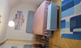 A modern bedroom with a large bed, an ottoman, and a rug in blue and beige tones. Decorative pictures are hanging on the wall, and there is a wardrobe.
