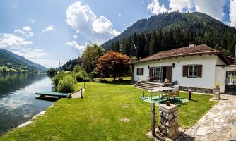 A beautiful garden with a modern villa by the lake. In the background, mountains and a clear sky are visible.