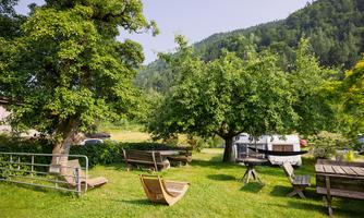 A green meadow with wooden benches and sun loungers under shade-giving trees. In the background, gentle hills can be seen.