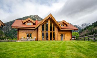 A beautiful wooden house with large windows in a green landscape. In the background, mountains and clouds can be seen.