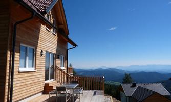 A modern wooden house with a terrace and a view of the mountains. The sky is clear and blue, creating a calm atmosphere.