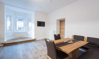 A modern dining room with a wooden dining table and comfortable chairs. The room has large windows and a television on the wall.