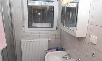 A small bathroom with gray tiles and a shower. There is a sink with a mirror and a window that lets daylight shine in.