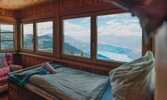 A cozy bedroom with a large window that offers a stunning view of the mountains and a lake. The room is made of wood, with a bed and a comfortable seating area.