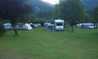 A campsite with motorhomes and tents surrounded by trees. The mountains are visible in the background, creating a beautiful natural setting.
