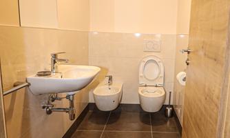 A modern bathroom with light walls and a dark tiled floor. There is a sink, a toilet, and a bidet visible.