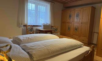 A cozy bedroom with two beds and large pillows. There is a wooden wardrobe and a table with chairs in front of a window.