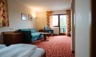 A cozy hotel room with blue sofas and a round table. Large windows with curtains let in plenty of light.