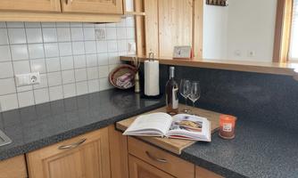 A modern kitchen with wooden cabinets and a dark work area. On the countertop are a cookbook, a bottle of wine, two wine glasses, and a candle.