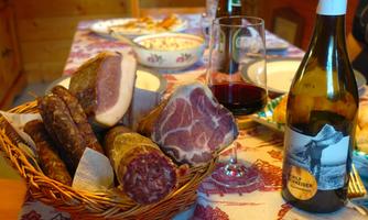 A basket with various types of sausages and ham is on a table. Next to the basket, there is a glass of red wine and a bottle of white wine.