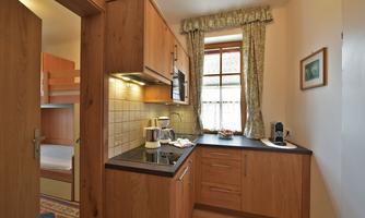A modern wooden kitchen with a cooking area and windows. In the background, a bathroom and a bedroom are visible.