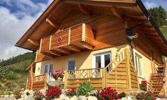 A beautiful wooden house with a balcony and colorful flower beds. In the background, gentle hills and blue sky can be seen.
