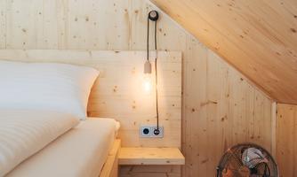 A cozy bedroom with wooden walls and a sloping roof. There is a large bed, a bedside lamp, and a fan.