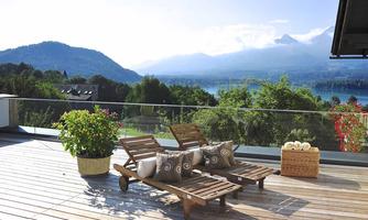 A terrace with two loungers and cushions, surrounded by plants. In the background, a picturesque landscape stretches out with mountains and a water lake.
