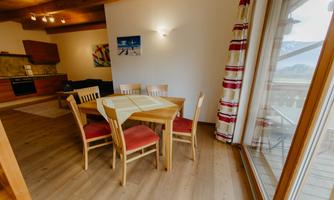 A brightly lit dining area with a wooden table and chairs. The room has large windows overlooking the surroundings.