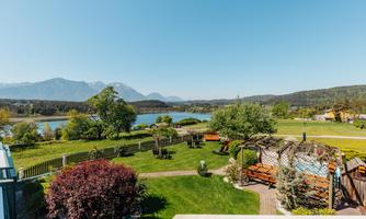 A picturesque landscape with a tranquil lake and gentle hills in the background. The garden is green and inviting, with seating areas and blooming plants.
