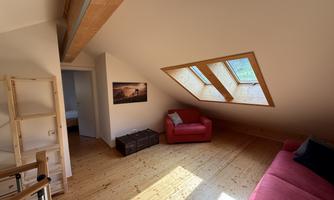 A cozy attic room with two windows that let in plenty of light. There is a red sofa and a small wooden table.