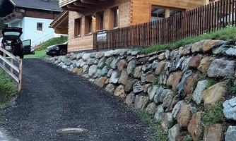 A cozy wooden house with a balcony, surrounded by a stone facade. The path to the house is lined with gravel and grass.