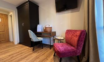 A cozy room with a red armchair and a desk. In the background, there is a TV and a wardrobe.
