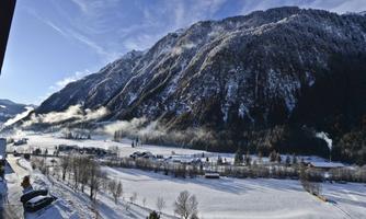 A snowy landscape with majestic mountains and a clear sky. The gentle hills and forests are covered in snow, and in the background, some houses can be seen.
