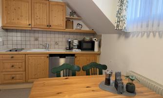 A cozy kitchen corner with wooden cabinets and a table. The equipment includes, among other things, a stove, a microwave, a dishwasher, and a coffee machine.