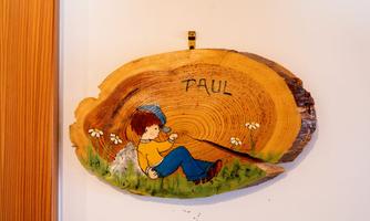 A wooden sign with the name "PAUL" and a colorful drawing of a boy sitting relaxed. Around him are flowers and a natural wood grain visible.