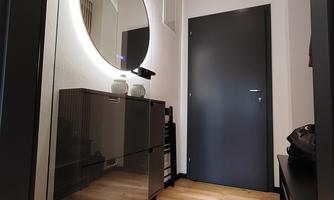 A modern hallway with a large, round mirror and a dark door. The floor is made of wood and there is a simple black cabinet.