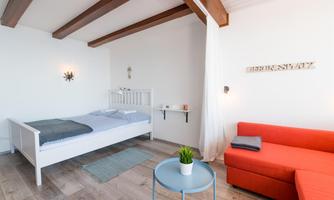 A modern bedroom with a single bed and a red sofa. The room has bright walls and wooden ceilings, as well as minimalist decoration.