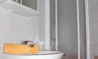 A modern bathroom with a sink and a shower cabin. On the sink, there are orange towels, and a colorful stool is standing next to it.