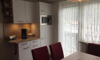 A modern kitchen with white cabinets and red chairs. The room has large windows with bright curtains and a pleasant light source.