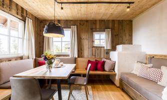 A cozy living room with wooden paneling and large windows. There is a dining table, seating, and a modern lamp.