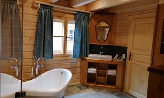 A cozy bathroom with a freestanding bathtub and wooden paneling. The windows are adorned with blue curtains and there is a wooden washbasin.