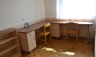 A bright room with a large window. There is a desk, two chairs, and a shelf.