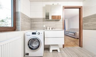 A modern laundry room with a washing machine, a white vanity, and a large mirror. In the background, an open living room is visible.