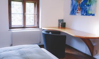 A modern bedroom with two large pillows and a desk. A colorful painting hangs on the wall and a window provides plenty of light.