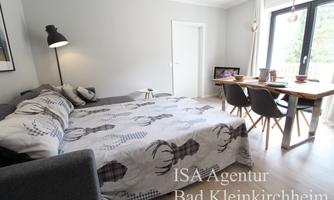 A modern room with a large bed, a dining table, and chairs. The walls are painted light, and there are windows that let in plenty of daylight.