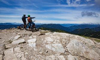 Two mountain bikers are standing on a rocky viewpoint, looking at the vast mountain landscape. The sky is clear with a few clouds.