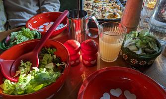 A table with colorful bowls, filled with fresh salad and a glass of various drinks. In the background, a casserole is visible.